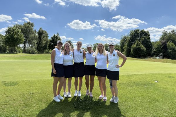 Sechs Frauen in weißem Poloshirt und dunklem Rock stehen auf einem Golfplatz zusammen und lächeln in die Kamera