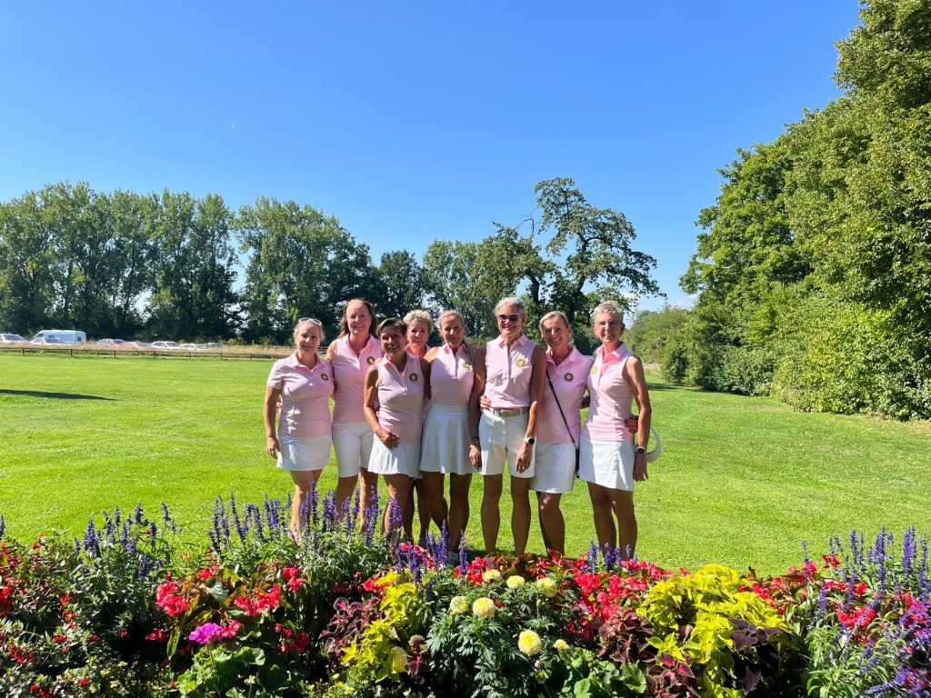 Sieben Frauen in rosa Poloshirts und weißen Röcken oder Shorts posieren lächelnd auf einem sonnigen Golfplatz. Vor ihnen befindet sich ein farbenprächtiges Blumenbeet, im Hintergrund stehen Bäume unter klarem blauem Himmel.