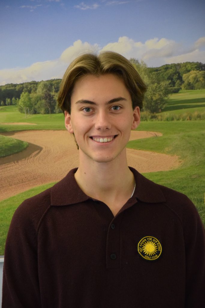 Junger Mann mit braunem Haar lächelt in die Kamera; er trägt einen dunkelbraunen Pullover mit einem Golf-Club-Abzeichen und steht vor einem Hintergrund mit einer Golfplatzlandschaft.