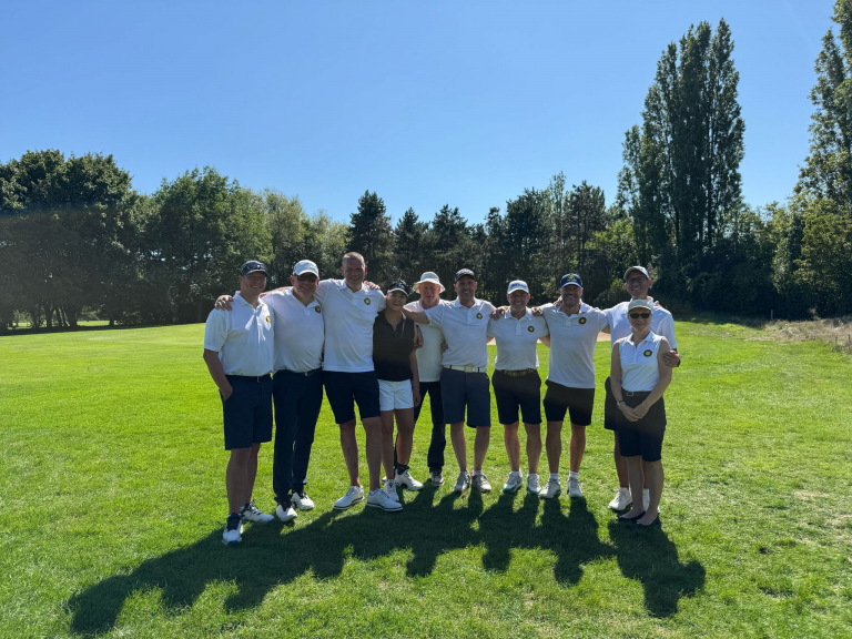 Eine Gruppe von zehn Menschen steht auf einem sonnigen Golfplatz nebeneinander, alle tragen sportliche Kleidung und lächeln in die Kamera.