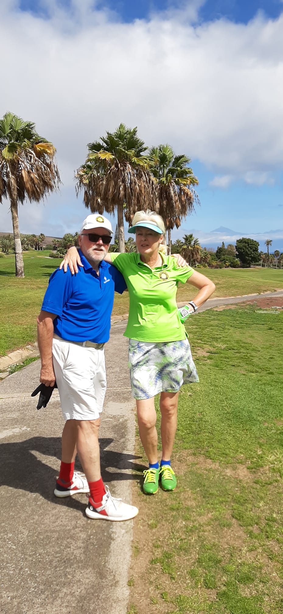 Zwei Personen stehen auf einem sonnigen Golfplatz zwischen Palmen. Beide tragen sportliche Golfkleidung und lächeln in die Kamera, während sie locker Arm in Arm posieren. Im Hintergrund sind Grünflächen, Bäume und ein leicht bewölkter Himmel zu sehen.