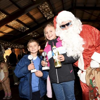 Zwei Kinder halten stolz ihre Pokale neben einer Person im Santa-Kostüm, die eine Geschenksack trägt. Die Szene findet in einer weihnachtlich dekorierten Halle statt.