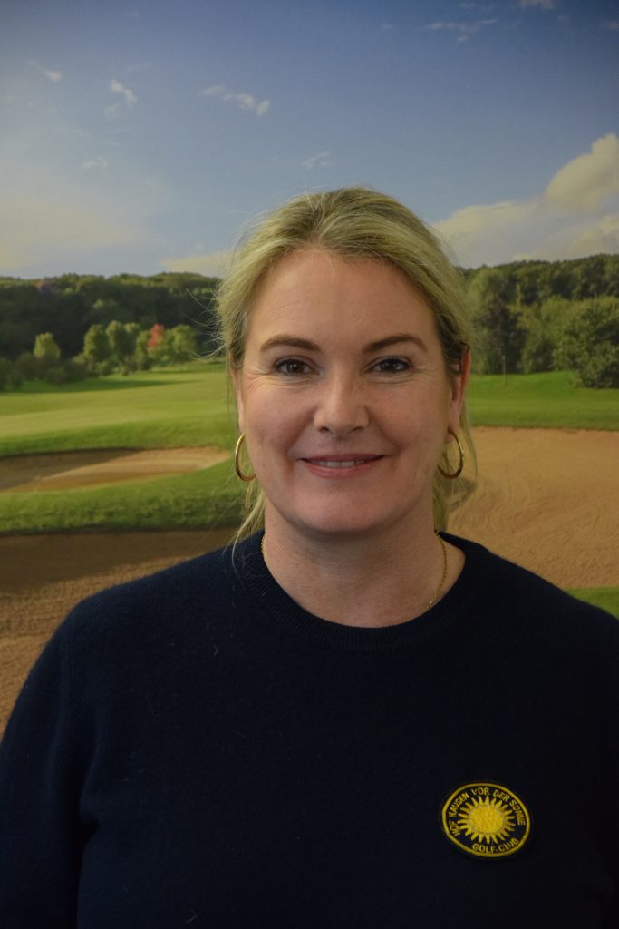 Eine Frau mit blondem Haar steht lächelnd vor einem Hintergrund, der eine grüne Golfanlage zeigt. Sie trägt einen dunkelblauen Pullover mit einem gelben Golfclub-Emblem.