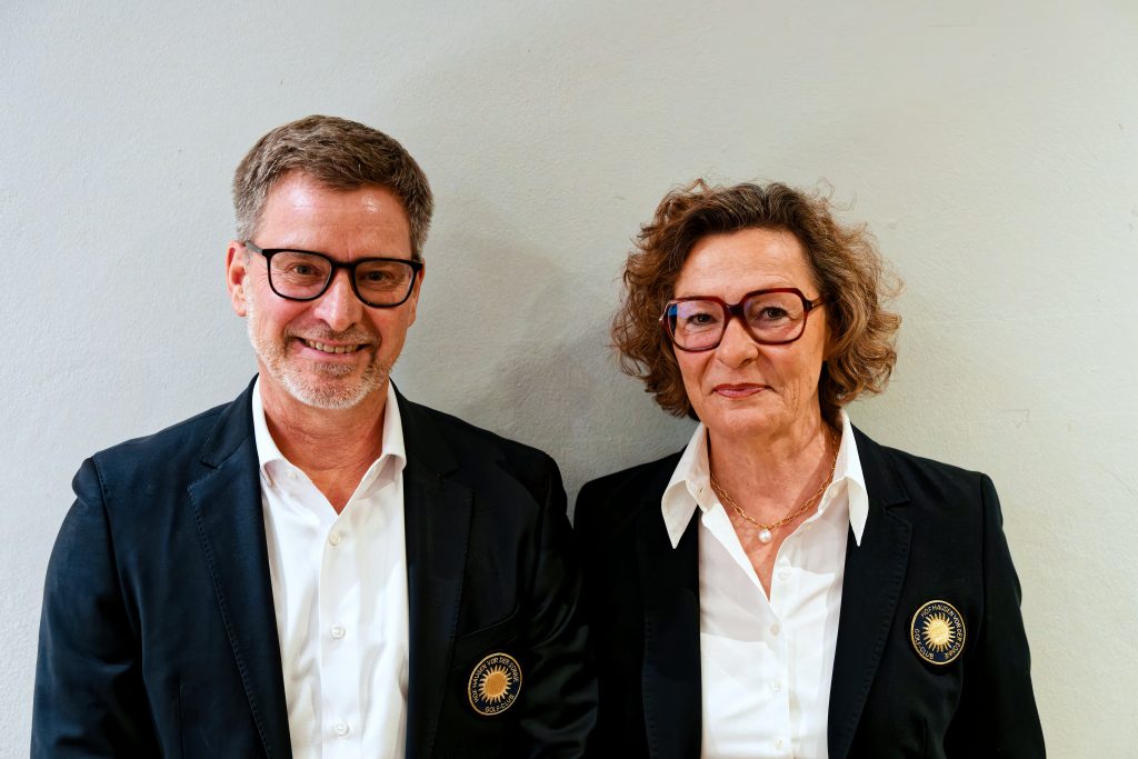 Zwei erwachsene Personen stehen nebeneinander vor einer hellen Wand. Beide tragen dunkle Blazer mit einem runden Abzeichen und weiße Hemden sowie Brillen und blicken lächelnd in die Kamera.