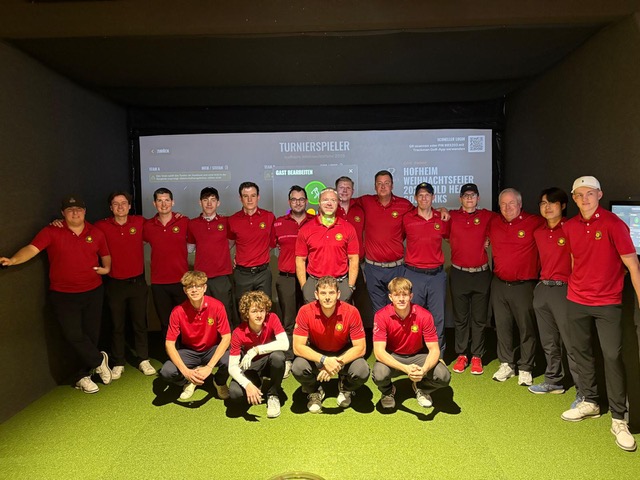 Gruppe von etwa 20 Personen in roten Poloshirts posiert gemeinsam in einer Indoor-Golfanlage vor einer Leinwand.