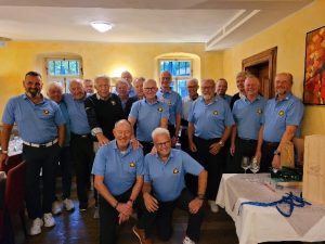 Gruppe älterer Männer in hellblauen Polo-Shirts steht und sitzt gemeinsam in einem hellen Raum und blickt lächelnd in die Kamera.