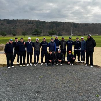 Gruppe von etwa 16 Personen in Sportkleidung posiert gemeinsam auf einem Golfplatz vor einer hügeligen Landschaft unter bewölktem Himmel.