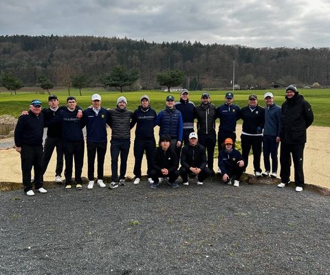 Gruppe von etwa 16 Personen in Sportkleidung posiert gemeinsam auf einem Golfplatz vor einer hügeligen Landschaft unter bewölktem Himmel.
