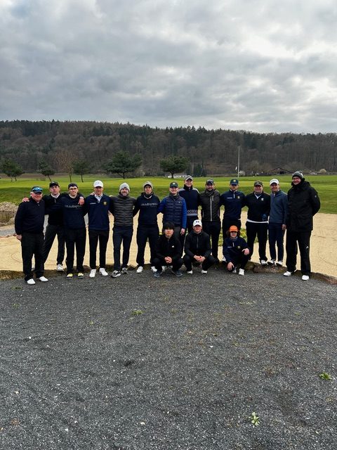 Gruppe von etwa 16 Personen in Sportkleidung posiert gemeinsam auf einem Golfplatz vor einer hügeligen Landschaft unter bewölktem Himmel.