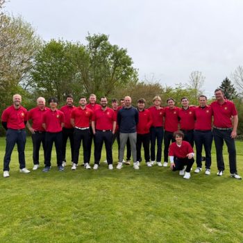 Gruppe von Männern in roten Poloshirts steht gemeinsam auf einer Wiese im Freien und blickt in die Kamera.