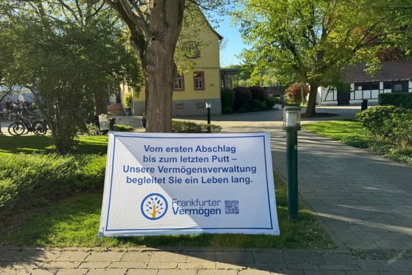 Werbebanner eines Vermögensverwalters auf einem gepflegten Gelände mit Bäumen und Gebäuden im Hintergrund.