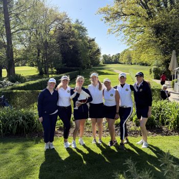 Eine Gruppe von sieben Frauen in Golfkleidung steht auf einem sonnigen Golfplatz zusammen; eine hält ein Baby. Im Hintergrund erstreckt sich eine grüne Fairway-Landschaft mit Bäumen und blauem Himmel.