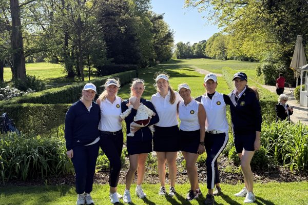 Eine Gruppe von sieben Frauen in Golfkleidung steht auf einem sonnigen Golfplatz zusammen; eine hält ein Baby. Im Hintergrund erstreckt sich eine grüne Fairway-Landschaft mit Bäumen und blauem Himmel.