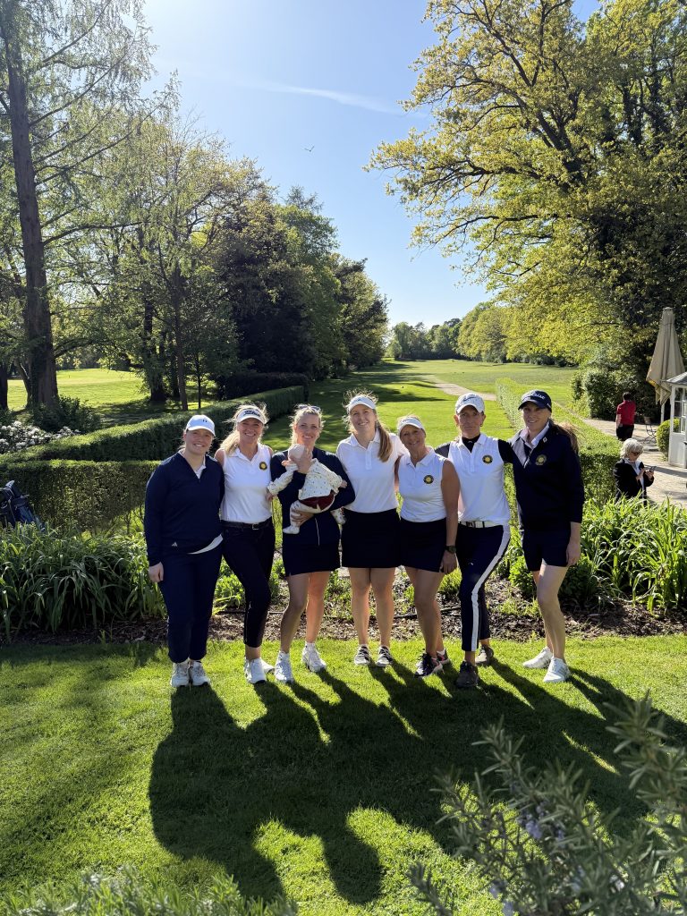 Eine Gruppe von sieben Frauen in Golfkleidung steht auf einem sonnigen Golfplatz zusammen; eine hält ein Baby. Im Hintergrund erstreckt sich eine grüne Fairway-Landschaft mit Bäumen und blauem Himmel.