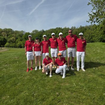 Acht Personen in roten Poloshirts und weißen Hosen posieren gemeinsam auf einem Golfplatz vor einer grünen Baumlandschaft bei sonnigem Wetter.