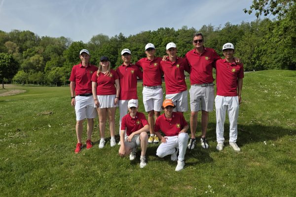 Acht Personen in roten Poloshirts und weißen Hosen posieren gemeinsam auf einem Golfplatz vor einer grünen Baumlandschaft bei sonnigem Wetter.