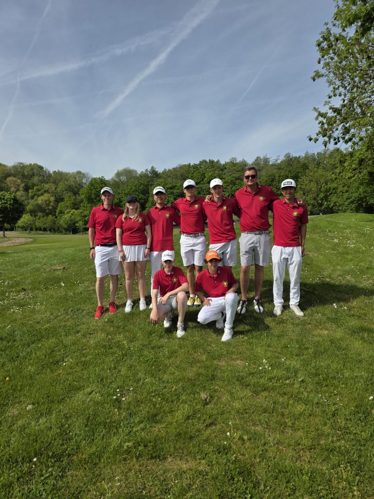 Acht Personen in roten Poloshirts und weißen Hosen posieren gemeinsam auf einem Golfplatz vor einer grünen Baumlandschaft bei sonnigem Wetter.