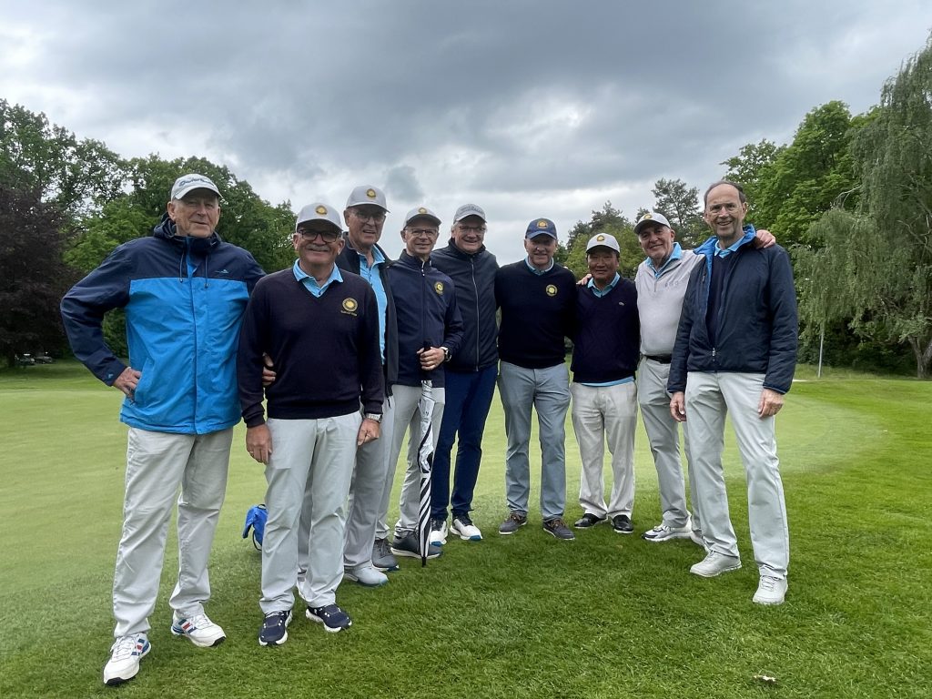 Neun Golfer in heller Sportkleidung stehen gemeinsam auf einem grünen Golfplatz unter dunklen Regenwolken und schauen in die Kamera.