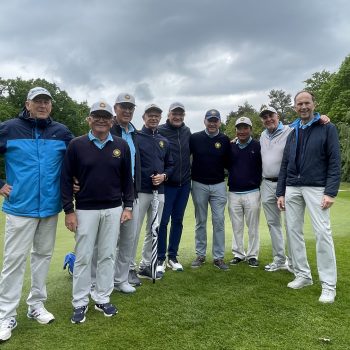 Neun Golfer in heller Sportkleidung stehen gemeinsam auf einem grünen Golfplatz unter dunklen Regenwolken und schauen in die Kamera.