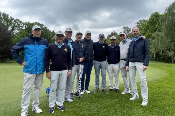 Neun Golfer in heller Sportkleidung stehen gemeinsam auf einem grünen Golfplatz unter dunklen Regenwolken und schauen in die Kamera.