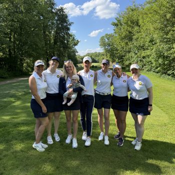 Sieben Frauen und ein Baby stehen gemeinsam auf einem sonnigen Golfplatz vor einer grünen Baumkulisse. Die Frauen tragen überwiegend weiße Poloshirts und dunkle Röcke oder Shorts. Die Gruppe lächelt in die Kamera, der Himmel ist blau mit wenigen Wolken.