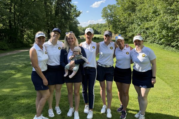Sieben Frauen und ein Baby stehen gemeinsam auf einem sonnigen Golfplatz vor einer grünen Baumkulisse. Die Frauen tragen überwiegend weiße Poloshirts und dunkle Röcke oder Shorts. Die Gruppe lächelt in die Kamera, der Himmel ist blau mit wenigen Wolken.