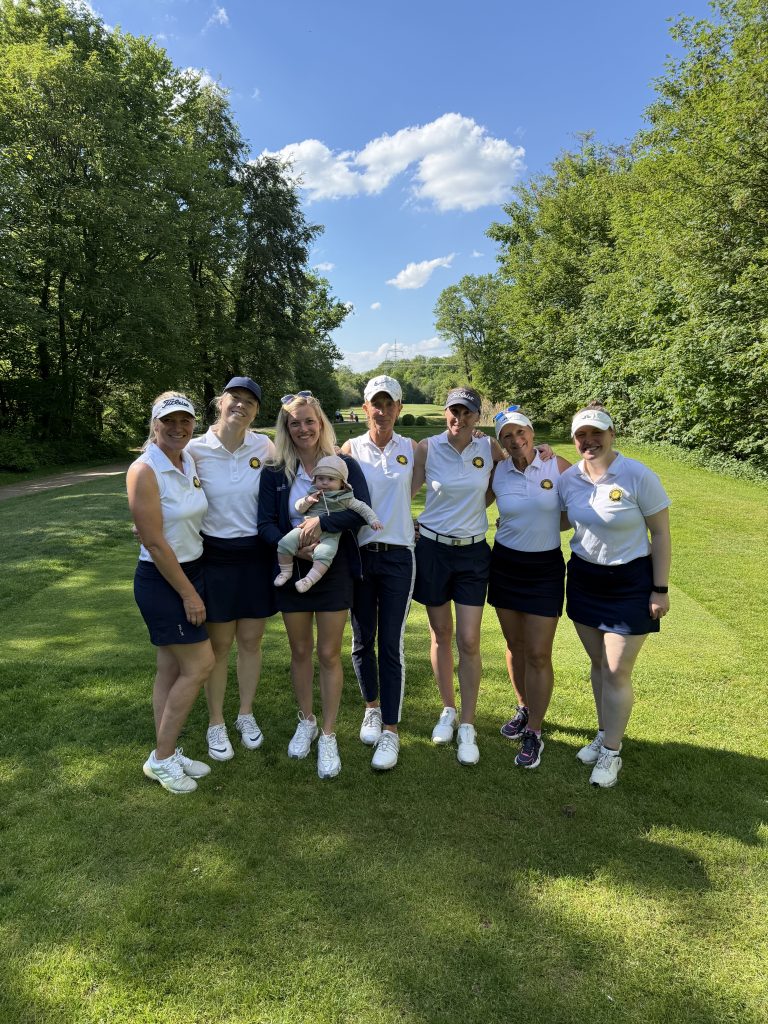 Sieben Frauen und ein Baby stehen gemeinsam auf einem sonnigen Golfplatz vor einer grünen Baumkulisse. Die Frauen tragen überwiegend weiße Poloshirts und dunkle Röcke oder Shorts. Die Gruppe lächelt in die Kamera, der Himmel ist blau mit wenigen Wolken.
