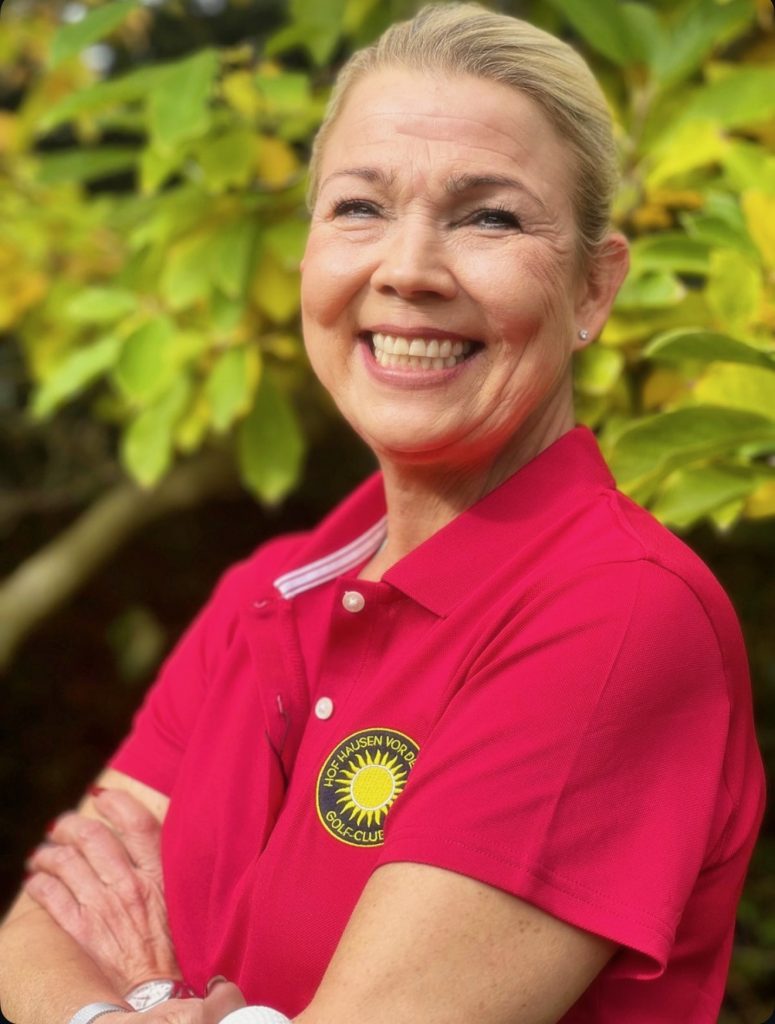 Lächelnde Frau in pinkem Golfclub-Poloshirt vor grünem Blätterhintergrund.