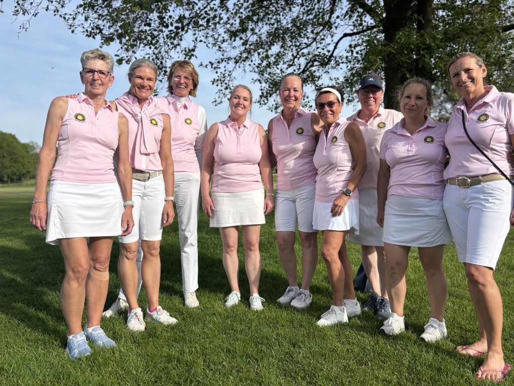 Gruppe von neun Frauen in einheitlichen rosa Poloshirts und weißen Golf-Outfits steht lächelnd auf einer Wiese unter einem Baum.
