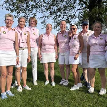 Gruppe von neun Frauen in einheitlichen rosa Poloshirts und weißen Golf-Outfits steht lächelnd auf einer Wiese unter einem Baum.