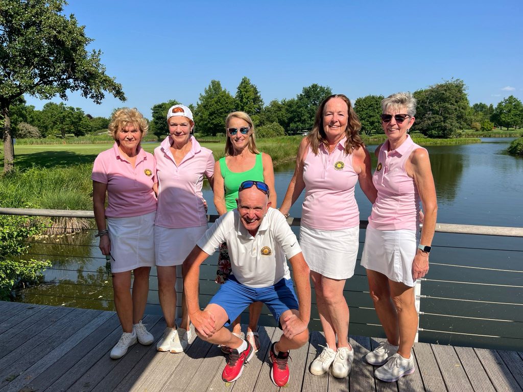 Sechs Personen posieren auf einer Brücke vor einem See in einer grünen Parklandschaft. Die meisten tragen rosa und weiße Sportkleidung, eine Person kniet vorne vor der Gruppe.