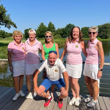 Sechs Personen posieren auf einer Brücke vor einem See in einer grünen Parklandschaft. Die meisten tragen rosa und weiße Sportkleidung, eine Person kniet vorne vor der Gruppe.