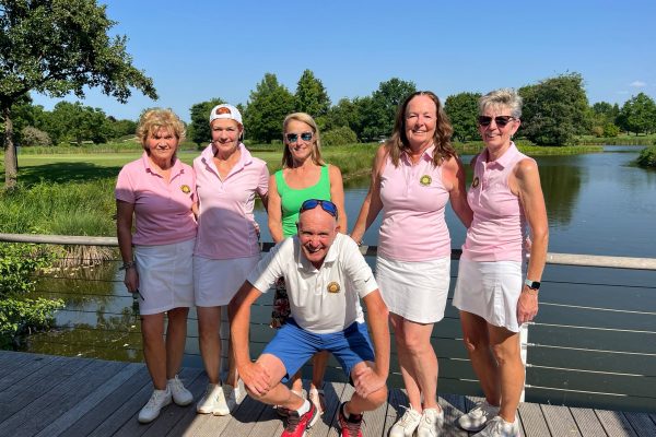 Sechs Personen posieren auf einer Brücke vor einem See in einer grünen Parklandschaft. Die meisten tragen rosa und weiße Sportkleidung, eine Person kniet vorne vor der Gruppe.