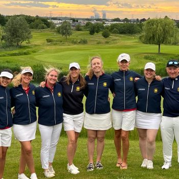 Acht Mitglieder einer Golfmannschaft stehen gemeinsam vor einem Wasserhindernis auf dem Golfplatz und posieren lächelnd in einheitlichen dunkelblauen Teamjacken und weißer Golfkleidung.