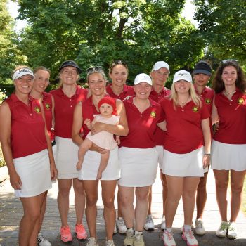 Die Damenmannschaft des Golf Club Hof Hausen vor der Sonne mit Trainer und Nachwuchs auf dem Golfplatz. Das Team trägt einheitliche rote Poloshirts mit Clublogo und weiße Röcke bzw. Hosen.