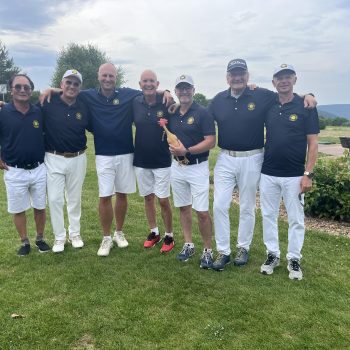 Sieben Mitglieder einer Herren-Golfmannschaft stehen gemeinsam auf dem Golfplatz und posieren lächelnd in einheitlichen dunkelblauen Poloshirts und weißer Golfkleidung.