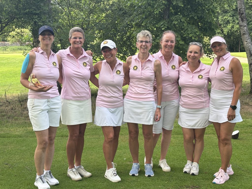 Sieben Damen der Golfmannschaft stehen gemeinsam auf dem Golfplatz und posieren lächelnd für ein Gruppenfoto in einheitlichen rosa Poloshirts und weißen Röcken beziehungsweise Shorts.