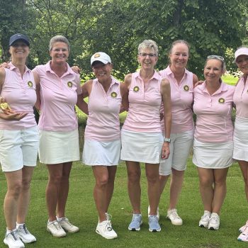 Sieben Damen der Golfmannschaft stehen gemeinsam auf dem Golfplatz und posieren lächelnd für ein Gruppenfoto in einheitlichen rosa Poloshirts und weißen Röcken beziehungsweise Shorts.
