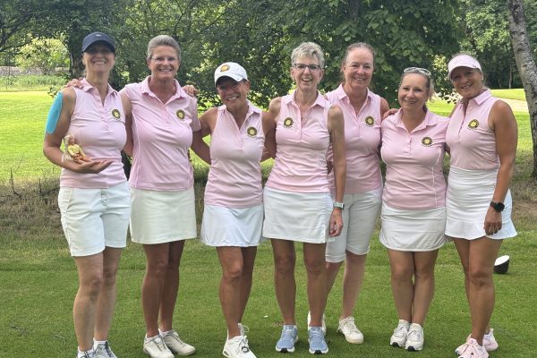 Sieben Damen der Golfmannschaft stehen gemeinsam auf dem Golfplatz und posieren lächelnd für ein Gruppenfoto in einheitlichen rosa Poloshirts und weißen Röcken beziehungsweise Shorts.