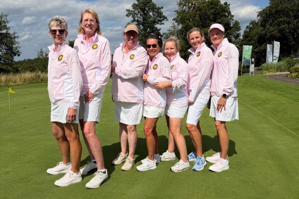 Die Damenmannschaft des Golf Club Hof Hausen vor der Sonne posiert gemeinsam auf dem Putting Green. Die Spielerinnen tragen einheitliche rosafarbene Teamjacken und weiße Röcke bzw. Shorts