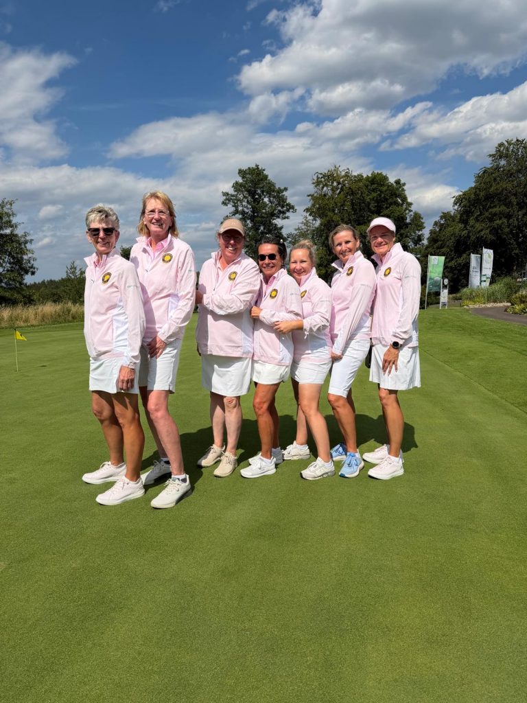 Die Damenmannschaft des Golf Club Hof Hausen vor der Sonne posiert gemeinsam auf dem Putting Green. Die Spielerinnen tragen einheitliche rosafarbene Teamjacken und weiße Röcke bzw. Shorts