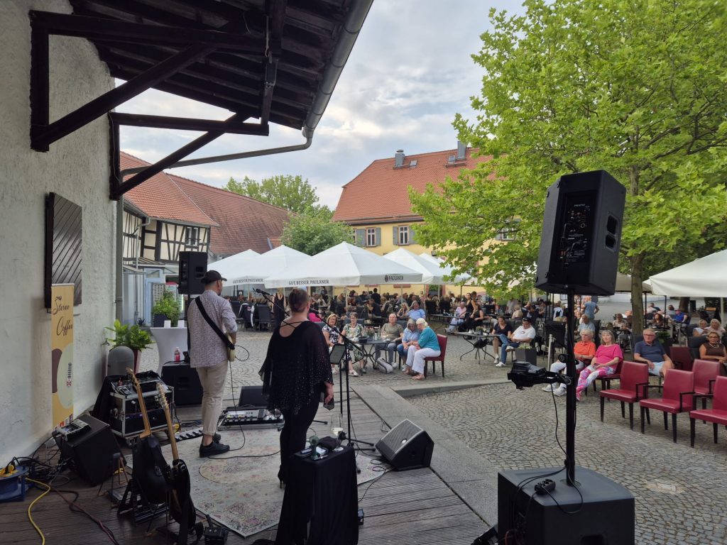 Die Band Stereo Coffee tritt auf einer Bühne auf der Hofinsel auf, während zahlreiche Gäste bei sommerlichem Wetter dem Konzert unter freiem Himmel lauschen.