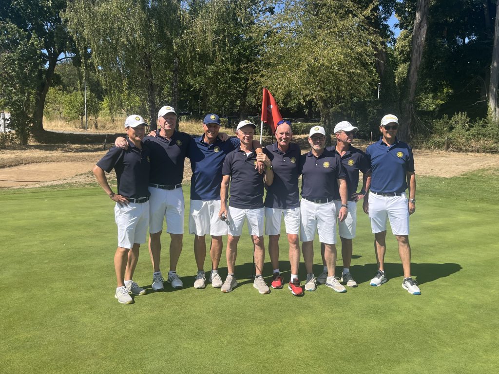 Acht Männer in Golfkleidung stehen gemeinsam auf einem sonnigen Golfplatz vor einem Grün. Hinter ihnen sind Bäume und zwei rote Fahnen zu sehen.