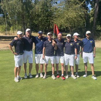 Acht Männer in Golfkleidung stehen gemeinsam auf einem sonnigen Golfplatz vor einem Grün. Hinter ihnen sind Bäume und zwei rote Fahnen zu sehen.
