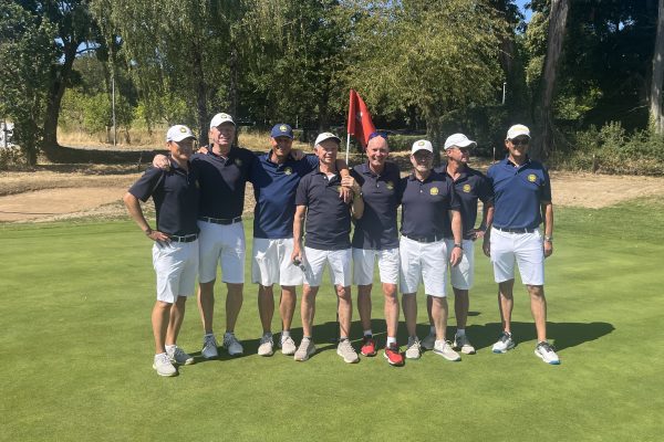 Acht Männer in Golfkleidung stehen gemeinsam auf einem sonnigen Golfplatz vor einem Grün. Hinter ihnen sind Bäume und zwei rote Fahnen zu sehen.