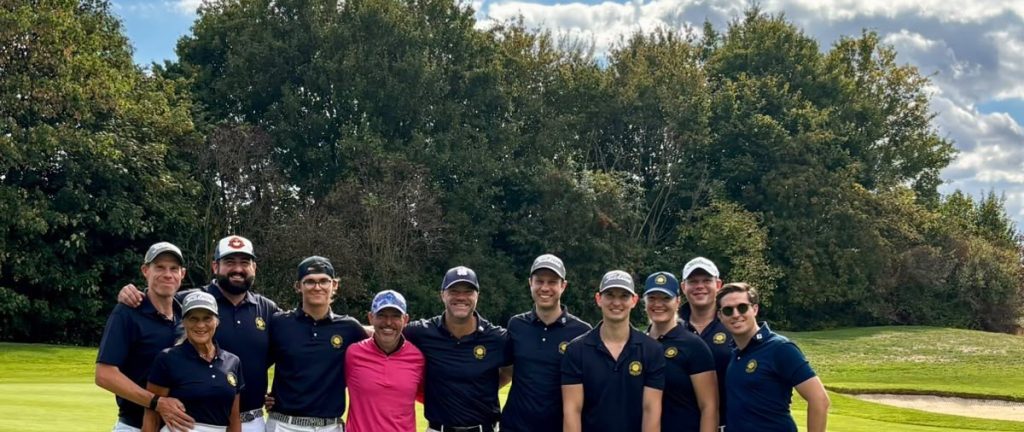 Mannschaftsfoto des Golfclubs Hof Hausen: zwölf Golferinnen und Golfer in dunkelblauen Clubshirts stehen lächelnd auf dem Grün vor hohen Bäumen.