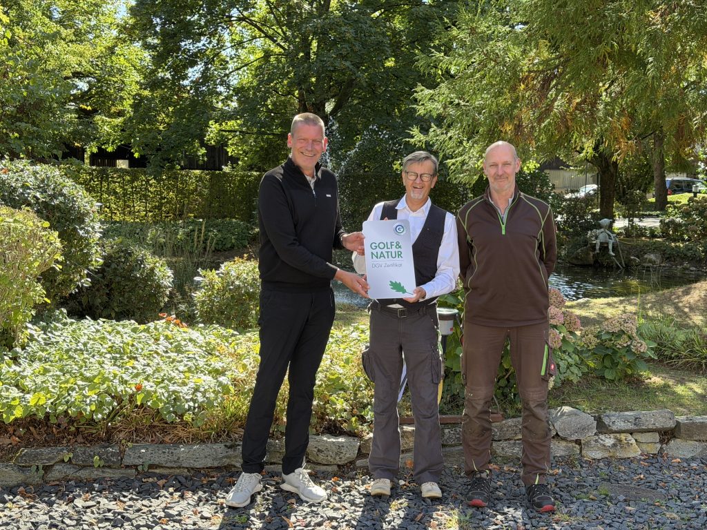 Drei Männer stehen vor einem Gartenbeet mit Teich und halten gemeinsam ein weißes Schild mit der Aufschrift „Golf&Natur DGV Zertifikat“ und einem grünen Eichenblatt.
