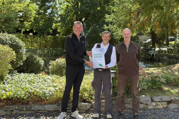Drei Männer stehen vor einem Gartenbeet mit Teich und halten gemeinsam ein weißes Schild mit der Aufschrift „Golf&Natur DGV Zertifikat“ und einem grünen Eichenblatt.