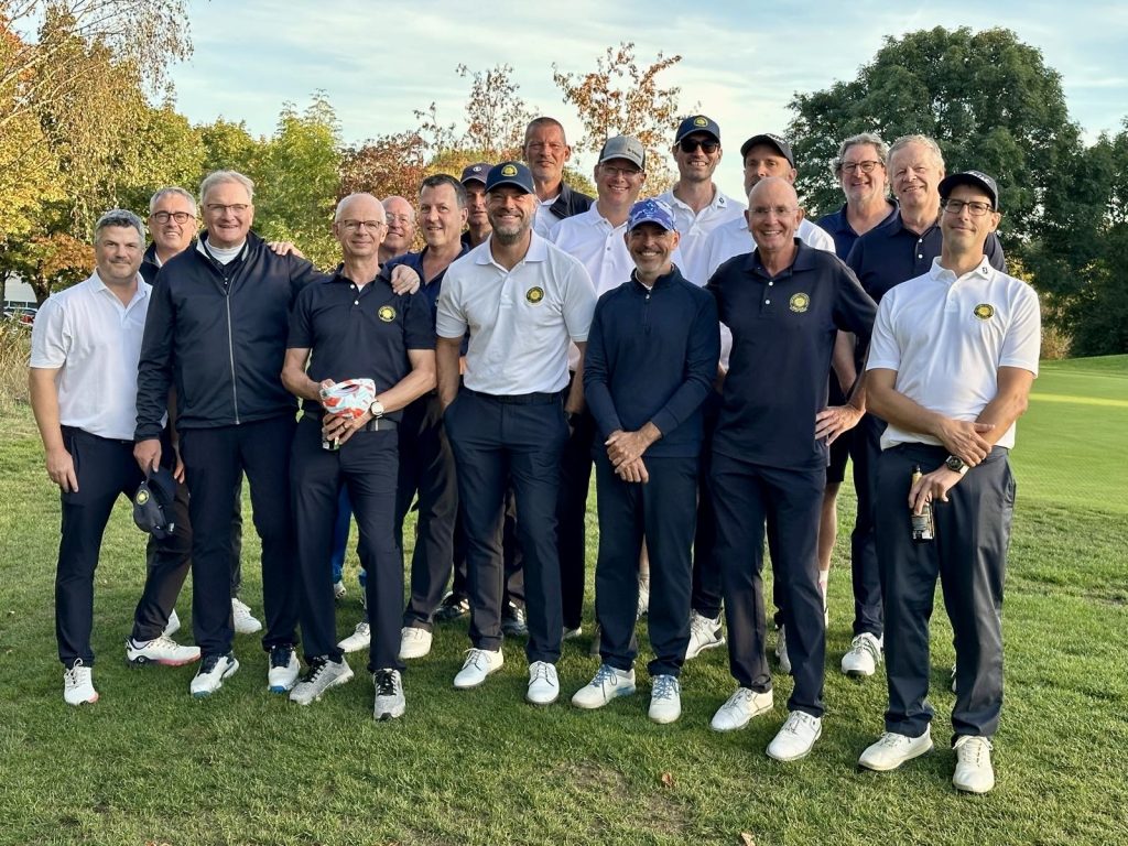 Gruppe von etwa zwanzig Golfern in weißen und dunkelblauen Clubpolos steht lächelnd auf dem Fairway vor herbstlichen Bäumen.