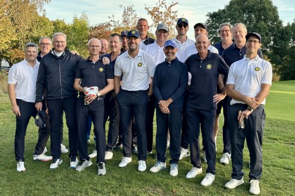 Gruppe von etwa zwanzig Golfern in weißen und dunkelblauen Clubpolos steht lächelnd auf dem Fairway vor herbstlichen Bäumen.